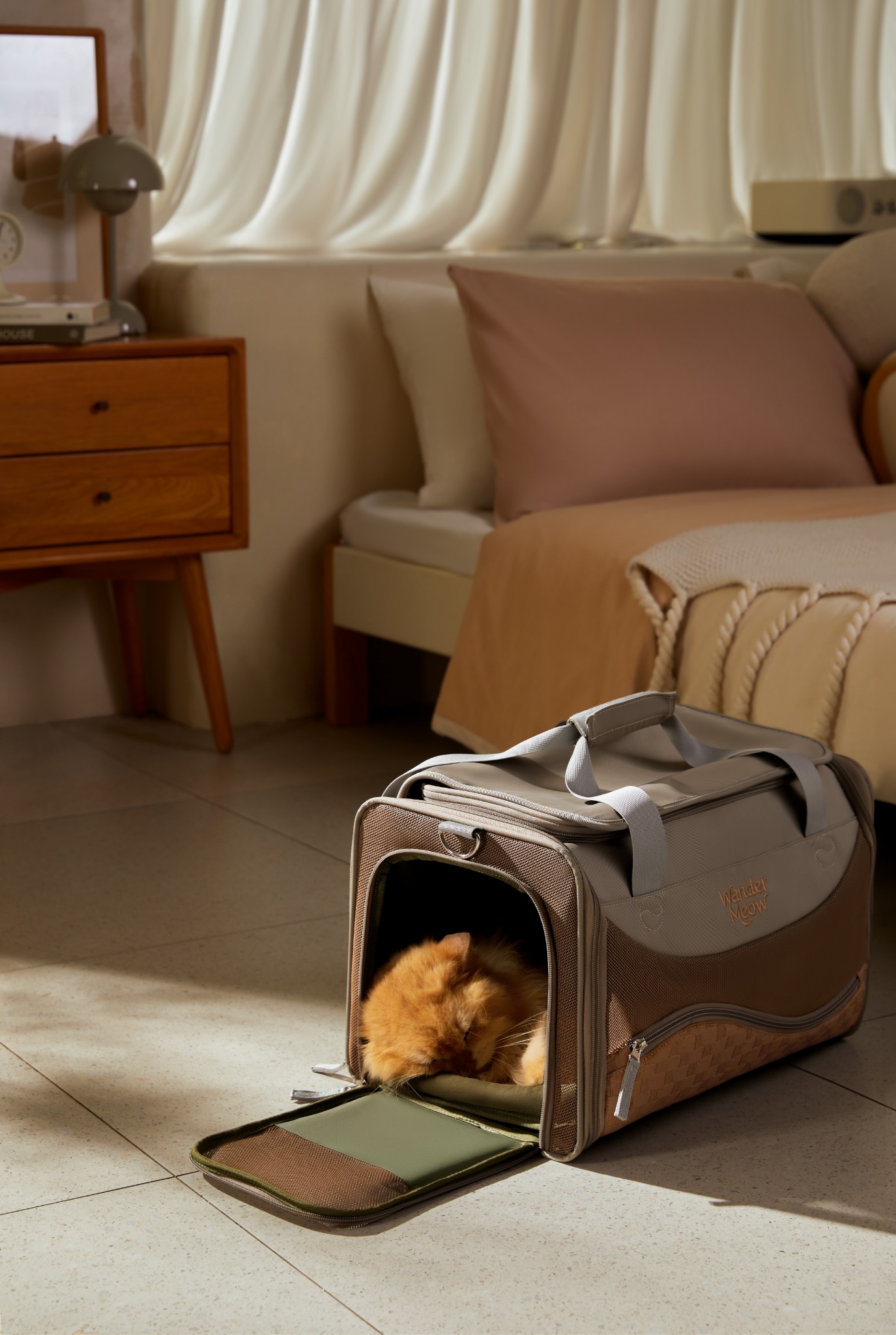 Orange cat resting at the open side doorway of the gray Wander Meow carrier in a sunlit room.